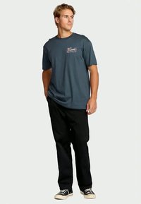 Jovem em t-shirt casual azul da Billabong, calças pretas e sapatilhas pretas, em pé, olhando para o lado com as mãos nos bolsos.