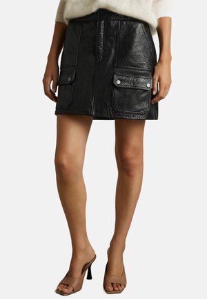 Femme portant une mini-jupe en cuir noir avec poches à rabat à l'avant, un pull beige et des sandales à talons hauts ouvertes marron.