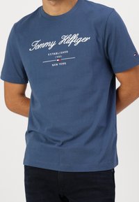 Kortärmad blå bomullströja med vit logotext som säger "Tommy Hilfiger" och året "1985" framtill, med rund halsringning.
