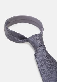 Cravat gri cu un model, realizată dintr-un material neted, având un design texturat cu un luciu subtil. Zona nodului este mai lată, oferind un aspect structurat.
