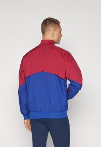 Röd och blå vindjacka med hög krage, elastisk fåll och kontrasterande färgblockdesign. Slät textur med synliga sömmar.