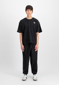 Ung mann står mot en enkel hvit bakgrunn iført en svart teksturert oversized t-skjorte og matchende bukser med hvite Adidas sneakers.