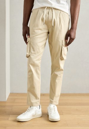 Pantalons cargo beiges avec coupe fuselée, poches latérales et taille élastique. Associés à des baskets blanches. Fond en bois.