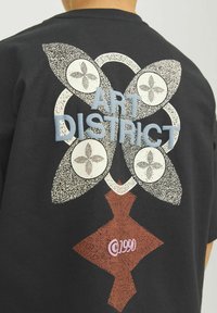 Svart t-shirt med en texturerad grafik med blommönster, texten "ART DISTRICT" och en röd diamantform. Designen innehåller olika mönster.