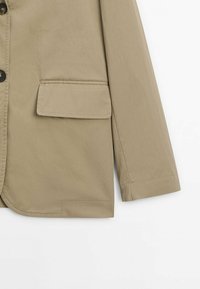 Veste beige avec une poche poitrine unique, une fermeture à boutons et une coupe ajustée. Tissu lisse avec des détails de couture minimaux et manches longues.