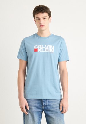 Lyseblå bomulds T-shirt med et hvidt og rødt grafisk design med "CALVIN KLEIN" tekst. Rund hals og korte ærmer.