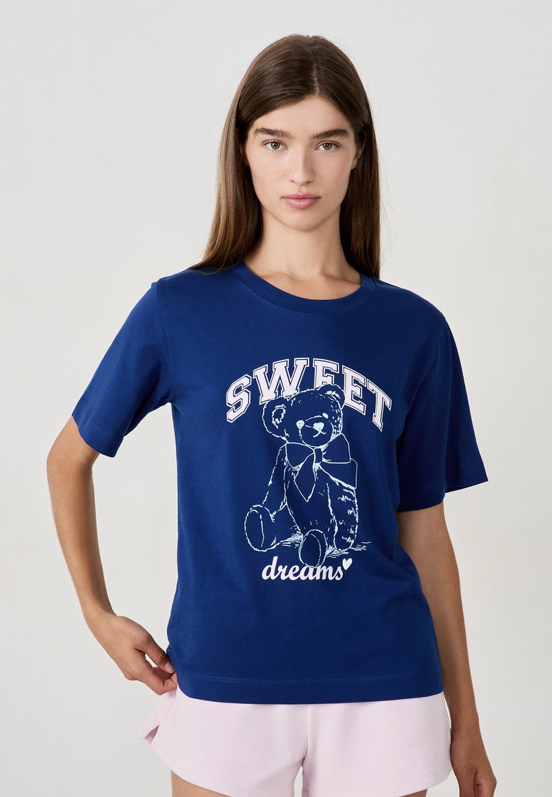 Femme aux cheveux bruns raides portant un t-shirt bleu avec un ours en peluche et le texte "SWEET dreams", associé à un short rose clair, fond uni.