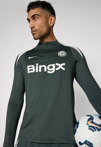 Cămașă sport cu mânecă lungă de culoare verde închis, cu accente albe și logo-ul "Bingx". Prezintă insigna Chelsea FC. Include o minge de fotbal cu un design patternat.