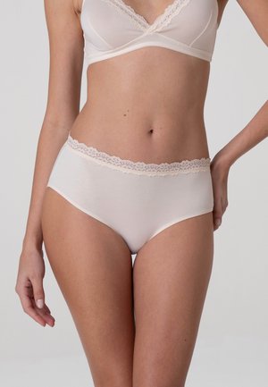 Donna che indossa biancheria intima color crema con rifiniture in pizzo e reggiseno abbinato su uno sfondo neutro e uniforme.