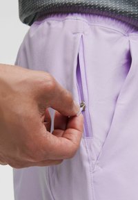 Shorts de sport violet clair avec une poche latérale zippée, dotés d'une texture lisse et d'une taille élastique. Main montrée en train d'utiliser le zip.