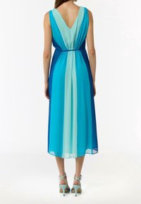 Femme portant une robe midi sans manches à dos en V avec des panneaux verticaux bleus et turquoise, associée à des sandales à talons hauts à fines lanières bleu clair.