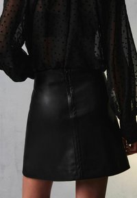 Gonna in pelle nera con un design aderente, caratterizzata da un orlo dritto e un sottile dettaglio di cucitura sul retro. Abbinata a una blusa trasparente nera a pois.