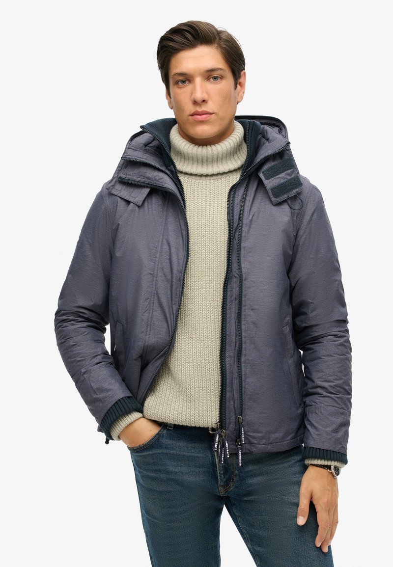 Mężczyzna w szarej kurtce z kapturem noszący kremowy sweter z golfem i niebieskie dżinsy, stojący z jedną ręką w kieszeni na białym tle.