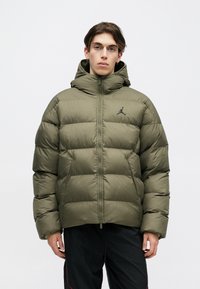Young man wearing an olive green puffer jacket with hood and black pants, standing against a plain light background.