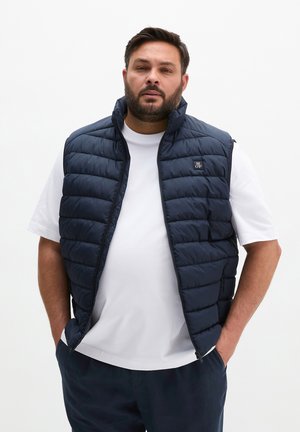 Mann trägt eine marineblaue gesteppte Weste über einem weißen T-Shirt mit den Händen in den Taschen vor einem einfarbigen weißen Hintergrund.