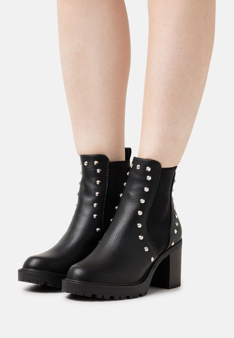 Bottines en cuir noir avec clous argentés et talons épais portées sur des jambes nues sur fond blanc.