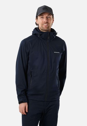 Man wearing a navy blue zip-up jacket with hood and black cap, standing against a plain white background, smiling slightly.