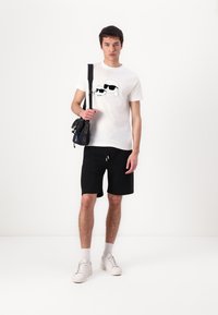 Maglietta bianca con design grafico nero, pantaloni corti neri e sneakers bianche. Gli accessori includono una piccola borsa a tracolla nera.