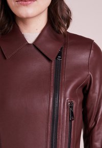 Burgunderrote Lederjacke mit spitzem Kragen, seitlichen Reißverschlüssen und einer glatten Textur, ergänzt durch zusätzliche Reißverschlusstaschen.