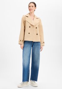 Beige zweireihige Jacke mit großen Knöpfen, weitem Revers und lockerem Schnitt, kombiniert mit hellblauen, weit geschnittenen Jeans und weißen Turnschuhen.