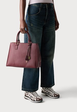 Bordeaux leren tote bag met ritssluiting, dubbele handvaten en een afneembare label. Model draagt een wijdvallende denim jeans en sportieve sneakers.