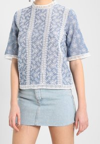 Light blue embroidered top with white floral patterns, short sleeves, and a high neckline. Features lace trim along edges. Paired with a denim skirt.