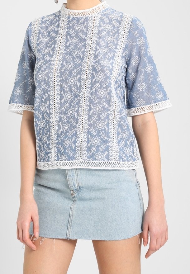 Light blue embroidered top with white floral patterns, short sleeves, and a high neckline. Features lace trim along edges. Paired with a denim skirt.