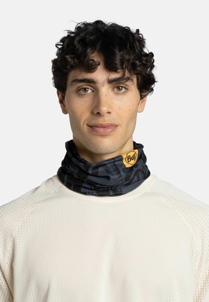 Joven con cabello oscuro y rizado que lleva una camisa blanca perforada y un protector de cuello negro con estampado y el logo amarillo de Buff.