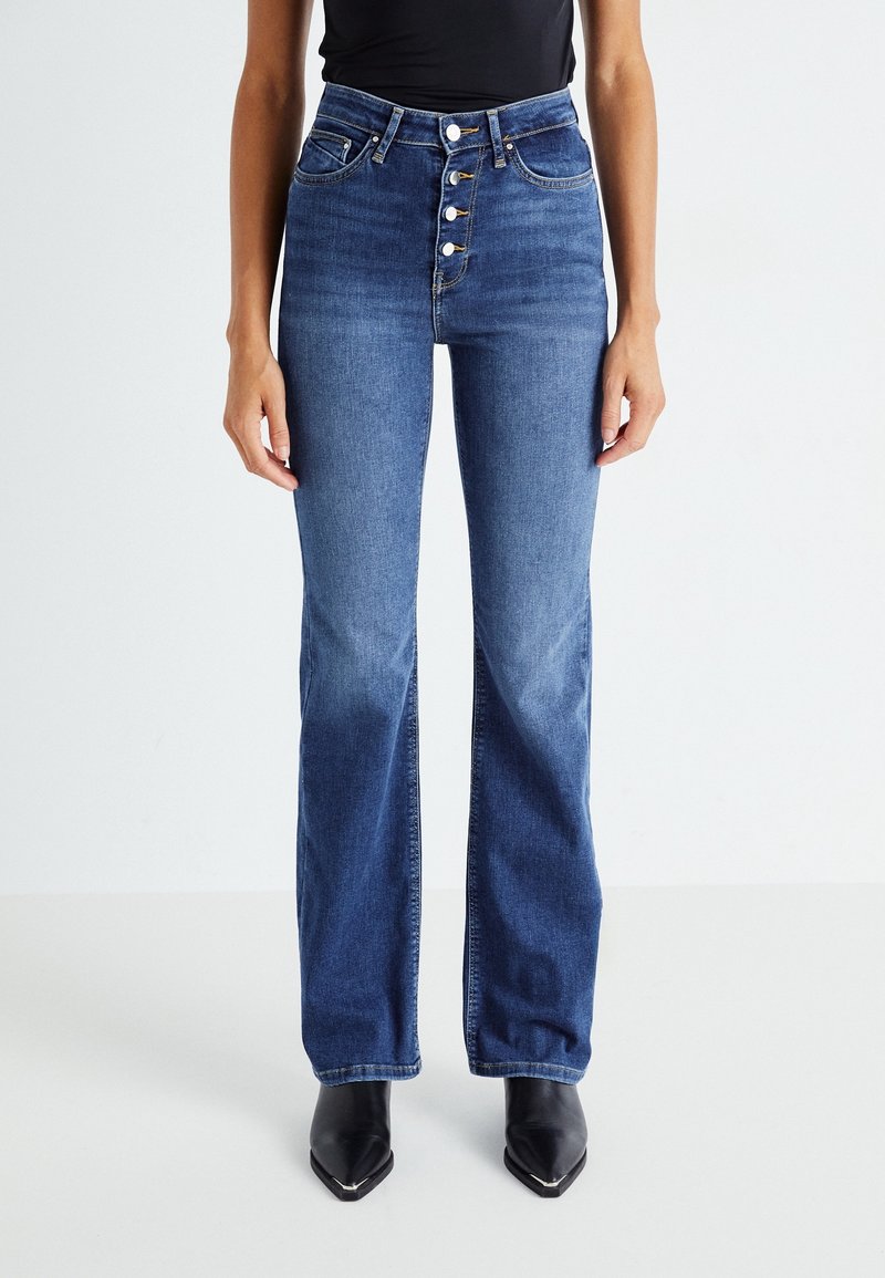 Jeans en denim bleu taille haute évasés avec fermeture à cinq boutons à l'avant, texture décolorée et poches latérales, associés à des chaussures noires à bout pointu.