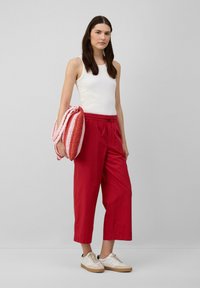 Pantaloni larghi rossi realizzati in tessuto leggero, abbinati a una canotta bianca. Tiene in mano una borsa intrecciata rosa e rossa, completata da sneakers beige.