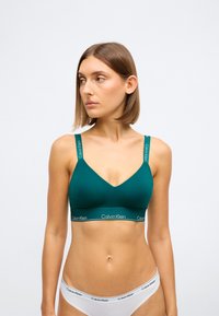 Teal Calvin Klein bralette s dizajnom u obliku v, širokim naramenicama i elastičnim pojasom s logotipom. Glatka tekstura tkanine i minimalni detalji.
