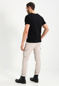Zwarte kortekous T-shirt gecombineerd met beige cargobroek met zijzakken en zwarte veterschoenen, gezien vanaf de achterkant.