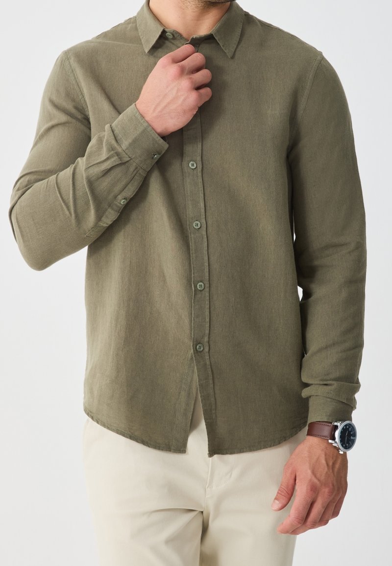 Homme portant une chemise vert olive boutonnée et un pantalon beige, ajustant le col avec la main gauche, portant une montre-bracelet marron au poignet droit.