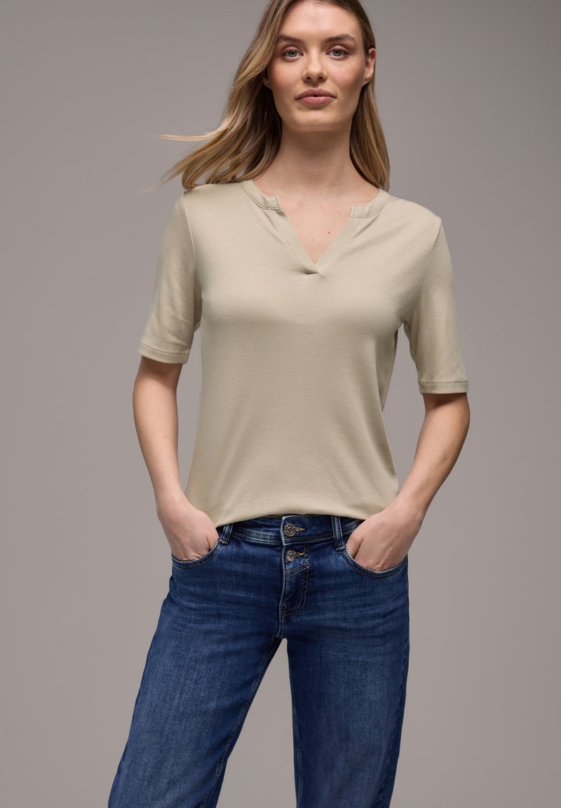 Junge Frau mit langen blonden Haaren, die ein beiges kurzärmliges Oberteil und blaue Jeans trägt, stehend mit den Händen in den Taschen vor grauem Hintergrund.