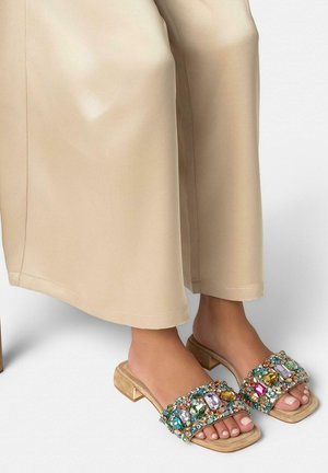 Pies con sandalias beige de tacón cuadrado decoradas con grandes gemas de colores, combinadas con pantalones beige de pierna ancha.