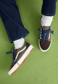 Des baskets marron et grises avec des lacets bleus, des accents blancs et des semelles en caoutchouc ; accompagnées d'un denim retroussé et de chaussettes blanches à côtes sur un fond vert.