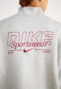 Grå sweatshirt med høy krage, burgunder brodert "NIKE Sportswear"-logo, "EST. 1972," og "JUST DO IT" under logoen.