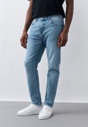 Ein Mann trägt hellblaue Jeans und weiße Turnschuhe, steht vor einem schlichten hellen Hintergrund, die Hände locker an den Seiten.