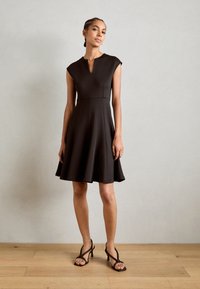 Musta fit-and-flare-mekko, jossa on hihaton malli, V-pääntie ja teksturoitu kangas. Mekossa on levenevä hame ja se on yhdistetty remmisandaaleihin.