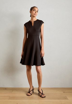 Schwarzes Fit-and-Flare-Kleid mit kurzen Ärmeln, V-Ausschnitt und strukturiertem Stoff. Es hat einen ausgestellten Rock und wird mit Riemchen-Heels getragen.