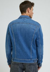 Mężczyzna zwrócony plecami, ubrany w niebieską dżinsową kurtkę i ciemnoszare dżinsy, na jednolitym jasnym tle.