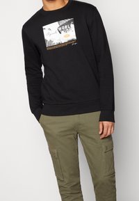 Felpa nera con maniche lunghe, caratterizzata da una stampa grafica di un panorama urbano e testo. Pantaloni cargo verde oliva con tasche laterali.
