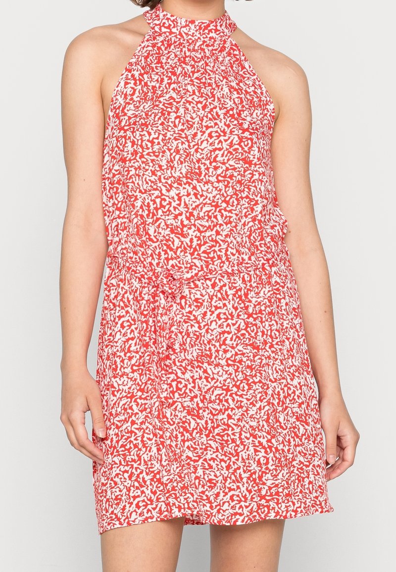 Femme portant une robe sans manches, longueur genoux, rouge et blanche à motifs, col licou avec ceinture à nouer, sur fond uni.
