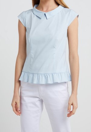 Femme portant un chemisier bleu clair à manches courtes avec un petit col et un ourlet à volants, assorti à un pantalon blanc, sur un fond uni.