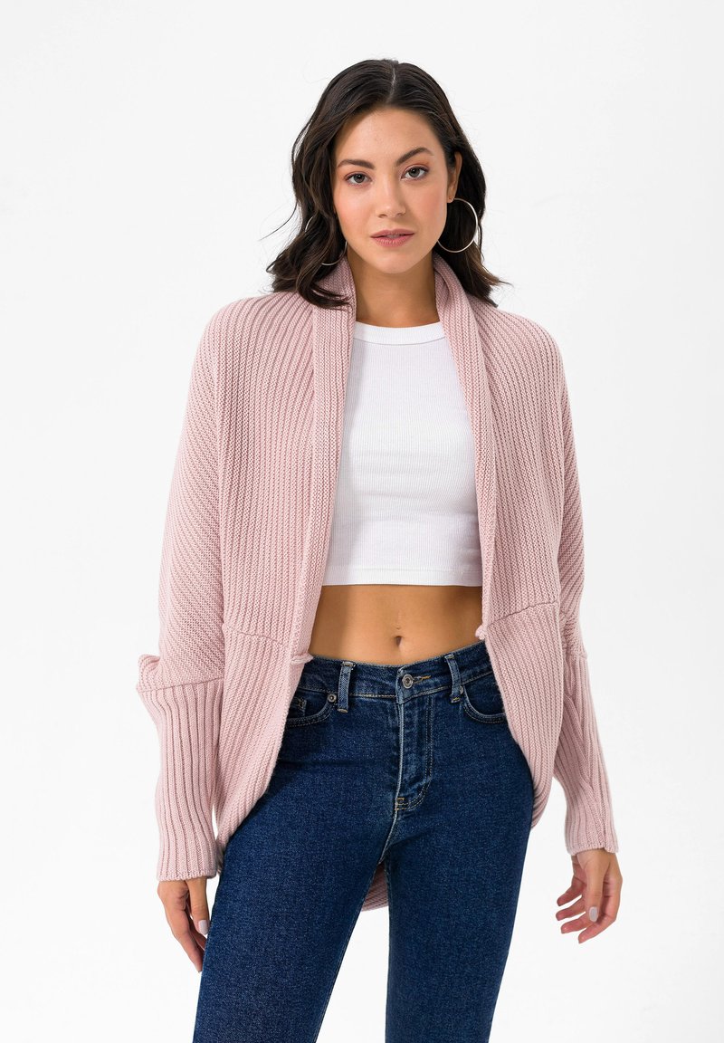 Hellrosa Strickjacke mit strukturiertem Rippenmuster, offener Front und langen Ärmeln, kombiniert mit einem weißen Croptop und blauen Jeans.