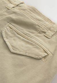 Gros plan sur un pantalon en tissu beige montrant une poche arrière avec rabat et passants de ceinture, mettant en valeur les coutures et les détails de la texture.