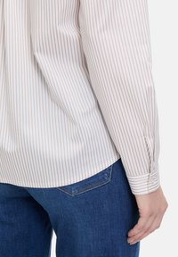 Long-sleeve shirt in white with beige vertical stripes, featuring a button cuff and a curved hem, paired with dark blue denim jeans.