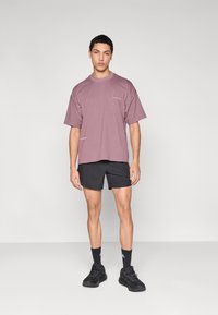 Κοντομάνικο, φαρδύ μπλουζάκι σε αποχρώσεις μοβ με τυπωμένες λεπτομέρειες, συνδυασμένο με μαύρο αθλητικό σορτς και μαύρα αθλητικά παπούτσια. Απλό φόντο.