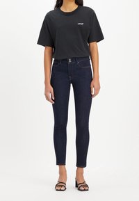 Camiseta de algodón negra de mangas cortas, con logo en el pecho, combinada con jeans ajustados de color azul oscuro y tacones negros con tiras.