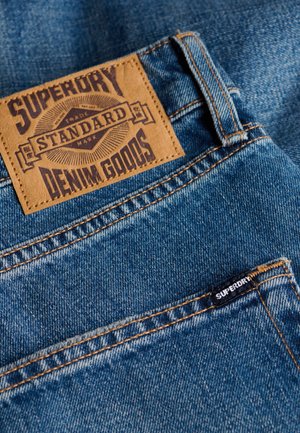 Jeans in denim blu con una toppa in pelle marrone con l'etichetta "SUPERDRY STANDARD DENIM GOODS" e una piccola etichetta nera sulla tasca.